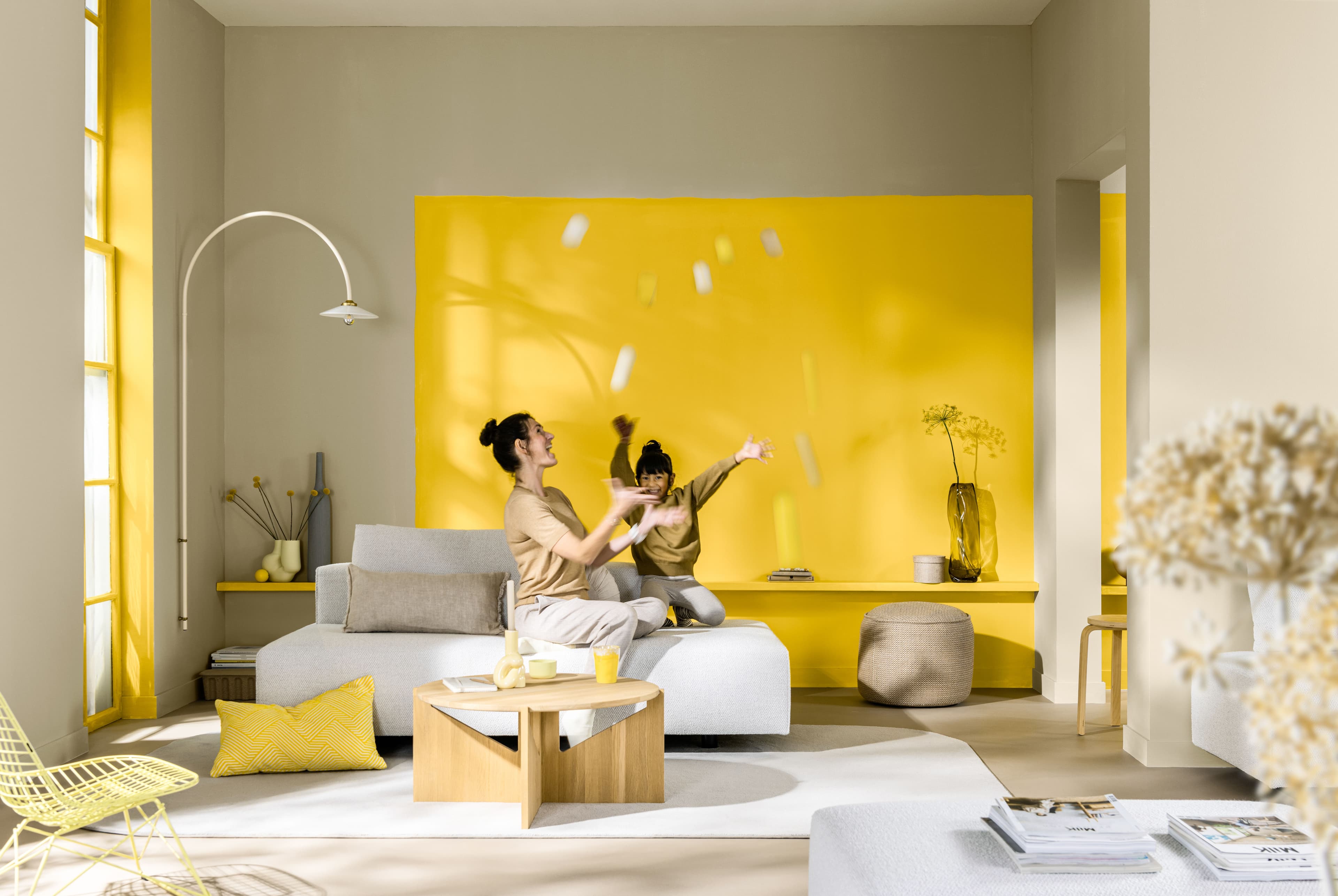 a young girl throws multi-coloured balls up in the air next to her mother in a living room. Behind them is a yellow statement wall.