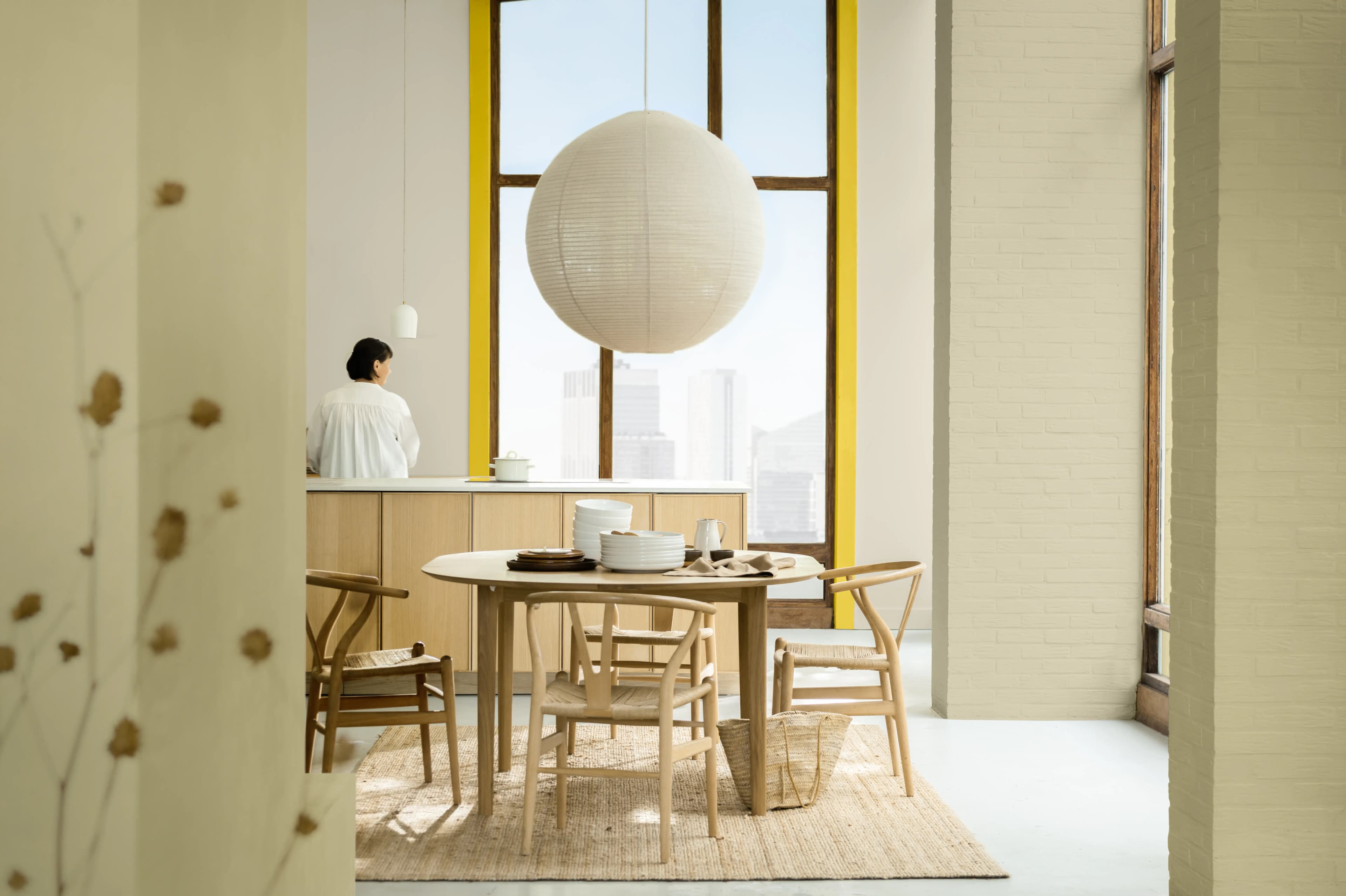A woman stands in an open-plan living room. A light hangs down from the ceiling and the window is framed with a lovely yellow.