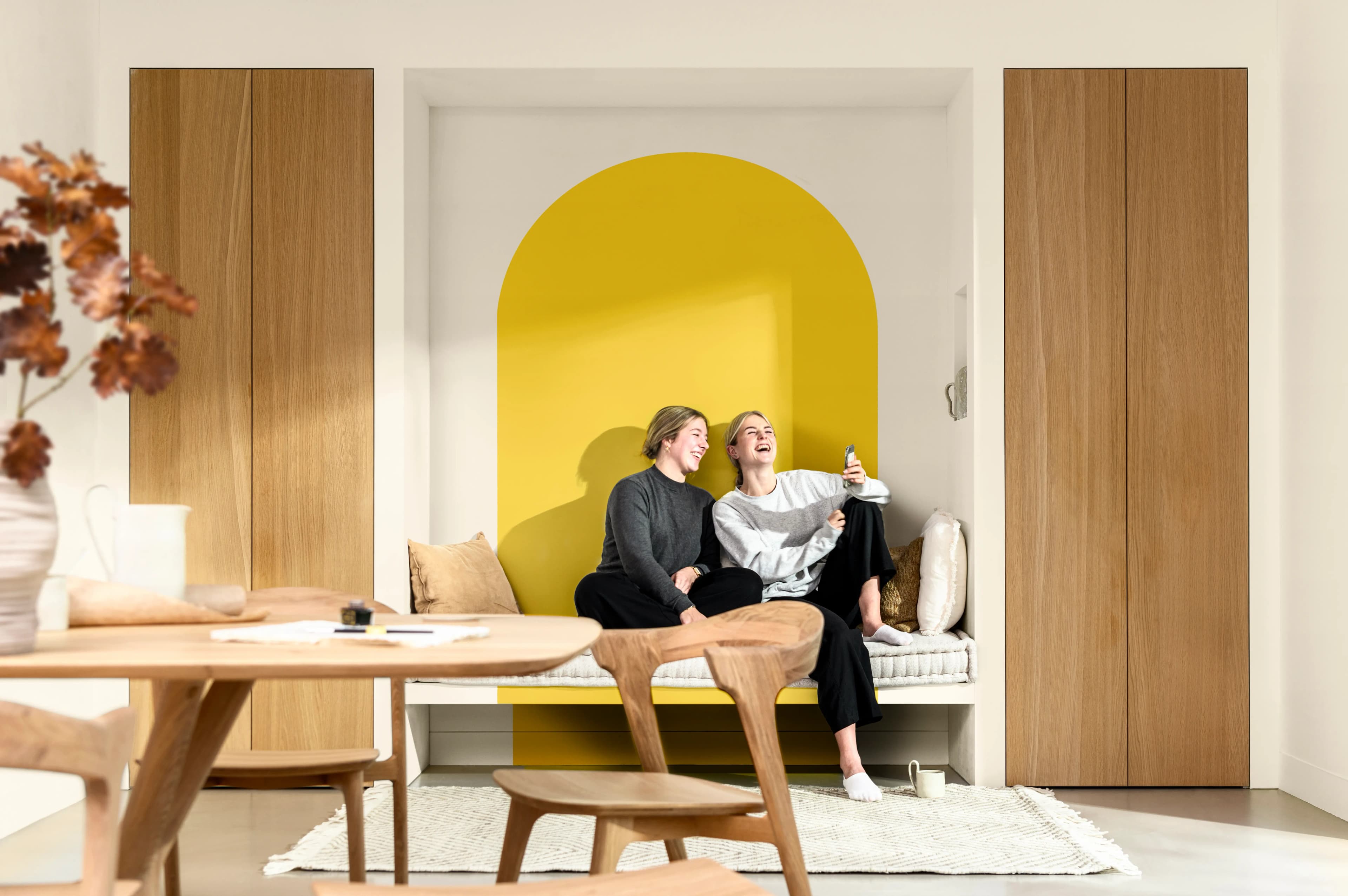 Two friends sit on a bench in a dining room. Behind them is an arch-shaped free-form shape in yellow.
