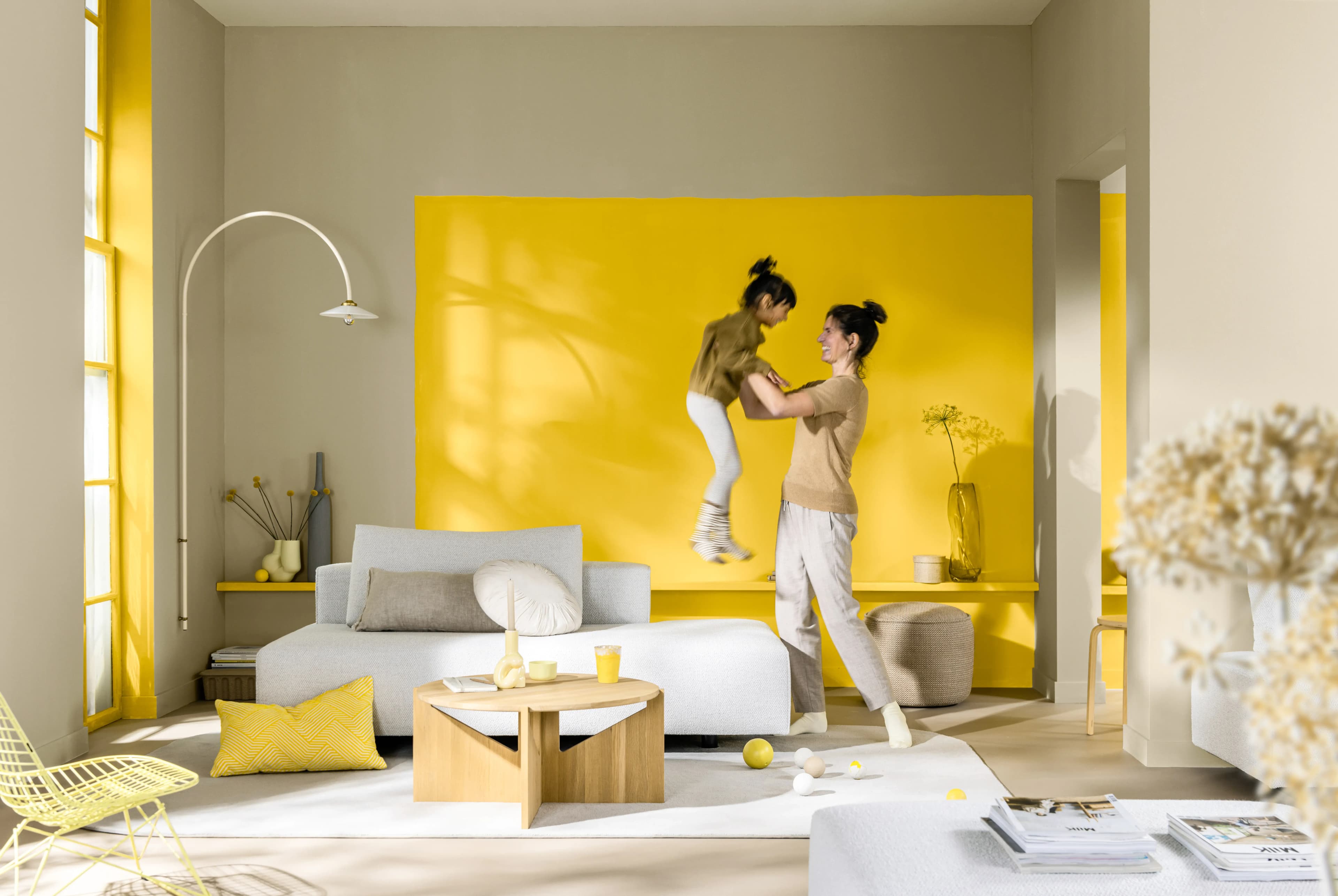 A parent lifting a child in a modern living room. Behind them is a statement feature wall painted in yellow.